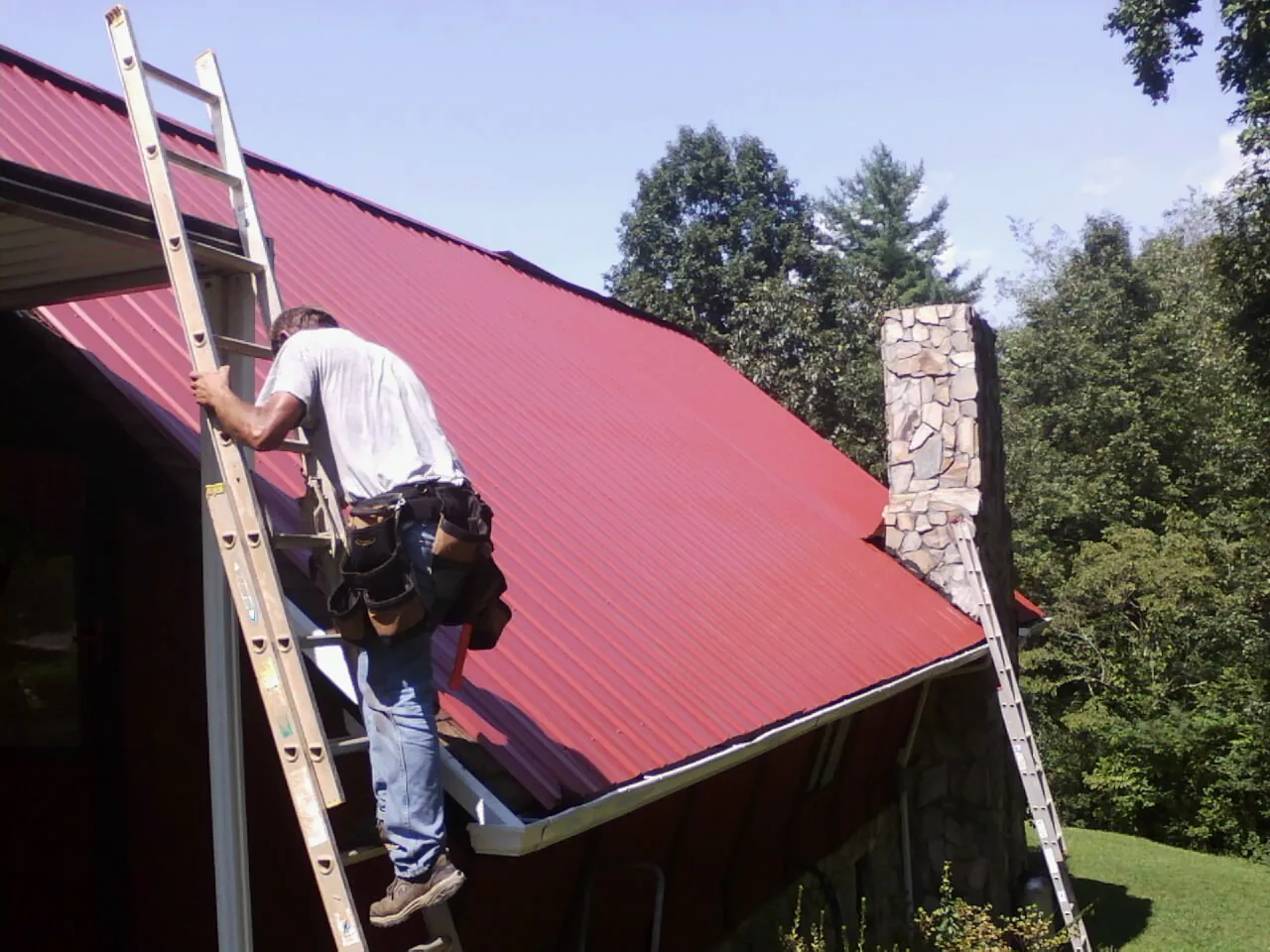 Roofer on a ladder beside a red metal roof and stone chimney — Soffit & Fascia Repair by Roofing Brothers in Wolfe City